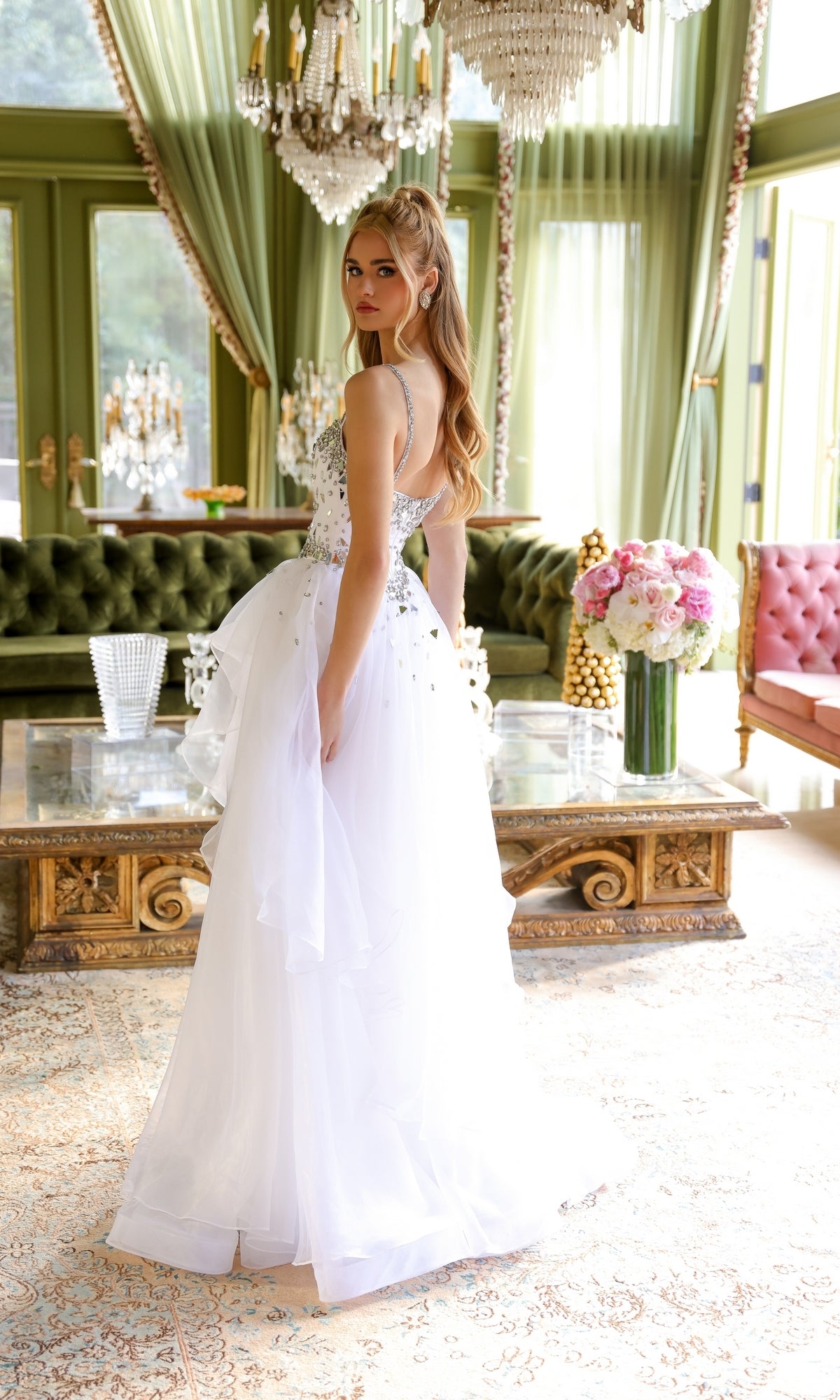 A woman in a white Tiered Long Prom Ball Gown, Ava Presley 29189, stands in an elegant room with green velvet sofas, chandeliers, and a large bouquet on a glass coffee table.